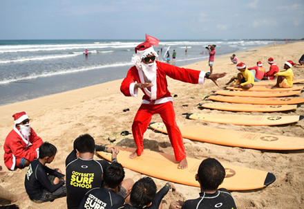 1 VĐV lướt ván trong bộ trang phục ông già Noel dạy trẻ em lướt sóng trên bãi biển Kuta trên đảo du lịch Bali của Indonesia.